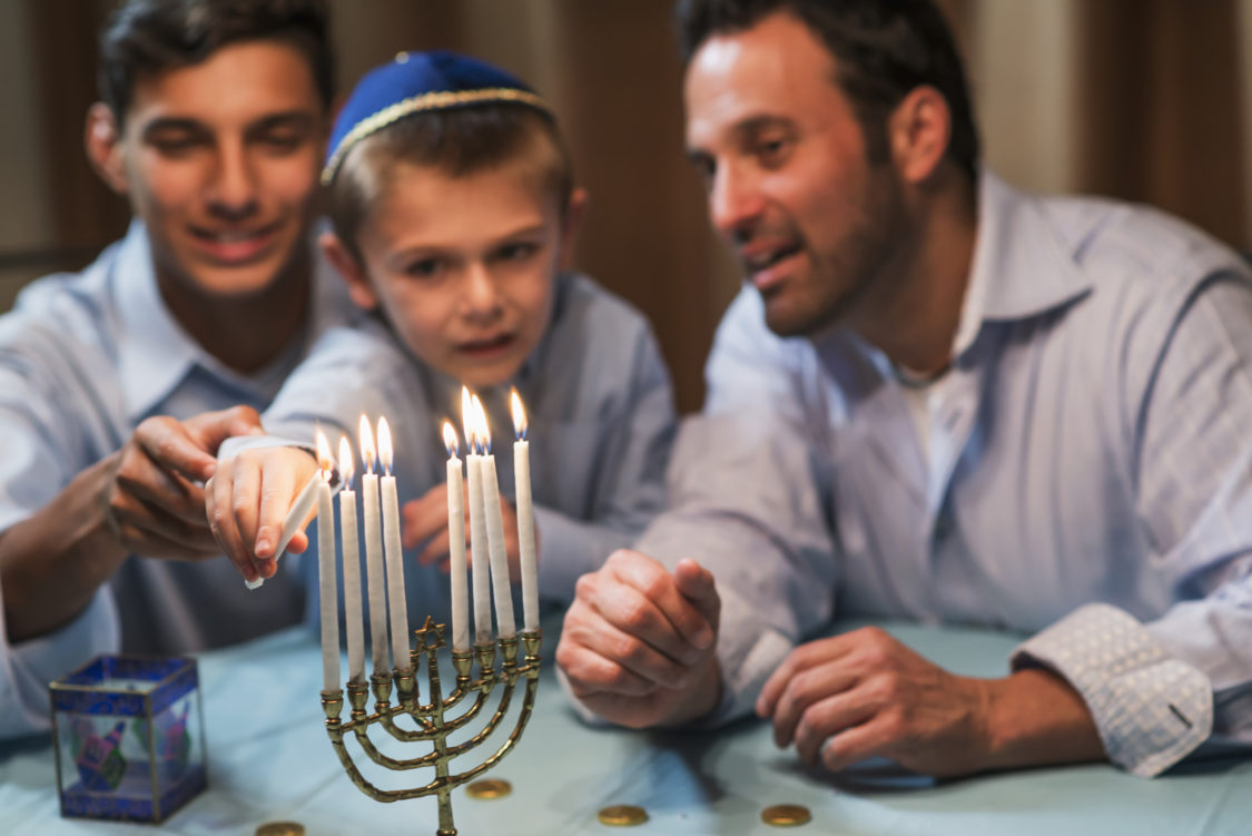 A Jewish father and his two sons celebrating hanukkah. The younger boy, 7 years old, is lighting a candle on the menorah. The focus is on the menorah.
