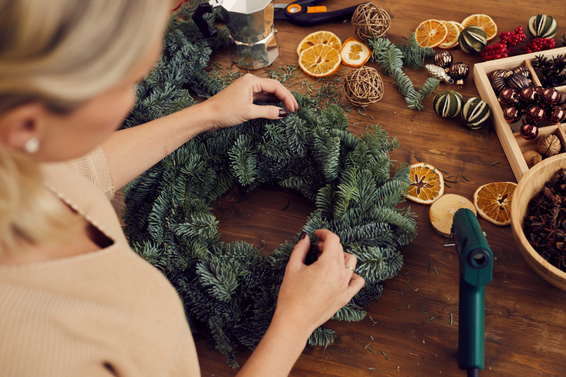 Vue en plongée d’une artisane debout à une table en bois pleine de décorations, en train de fabriquer une couronne de Noël dans un atelier.