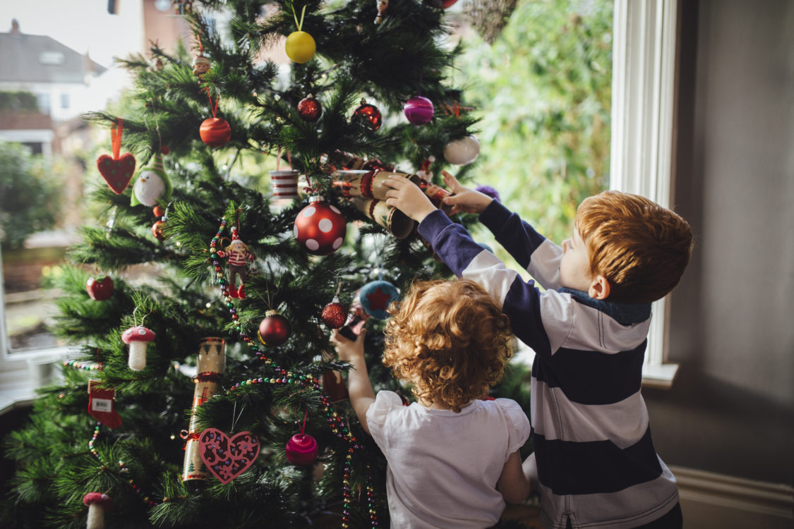 A young red head boy and his sister are decorating the Christmas tree in their home. He is reaching up and putting a bauble onto the tree while his sister tries to help.