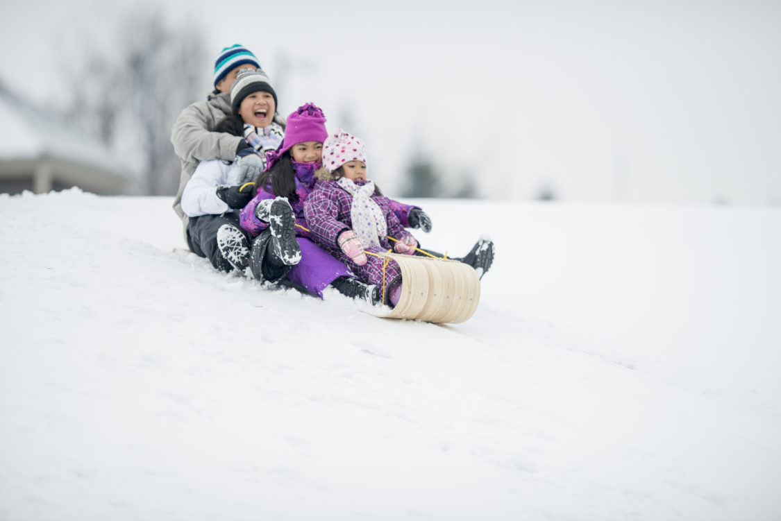 A family (father, mother, and daughters) enjoying winter by riding a toboggan sled/tube down a snowy hill in winter.