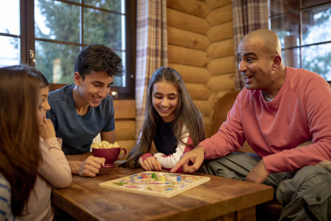 Une famille joue à un jeu de société ensemble dans une cabane en rondins. Un homme d'âge moyen sourit et montre du doigt le jeu tout en parlant à sa fille.