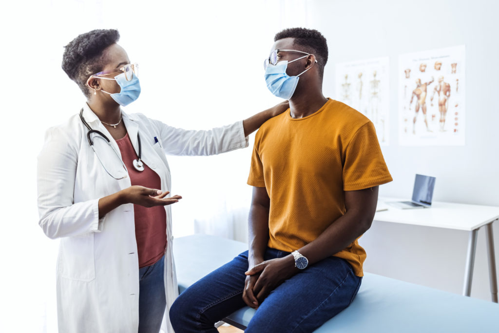 Femme médecin noire communiquant avec un patient tout en portant un masque protecteur lors d'un rendez-vous médical.