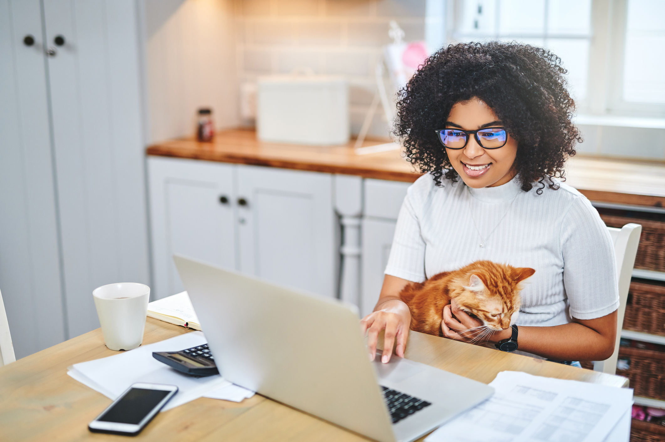 Photo of a young woman affectionately holding her cat and using a laptop while working from home