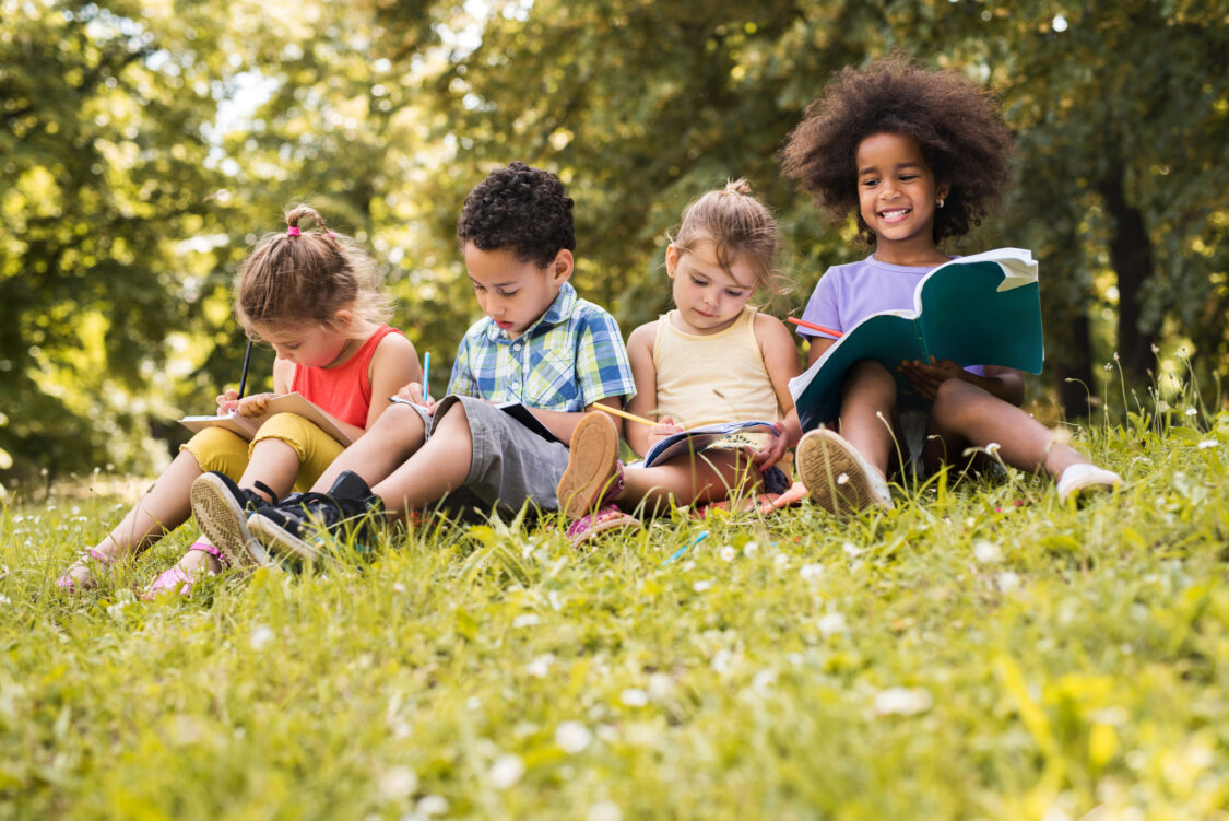 Quatre petits enfants assis dans l'herbe au parc et écrivant dans leurs cahiers.