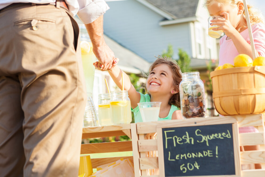 Cheerful little girl shakes hands with her first lemonade stand customer. A Caucasian man is standing in front of the stand. A jar full of coins and a basket of fresh lemons is on the stand.