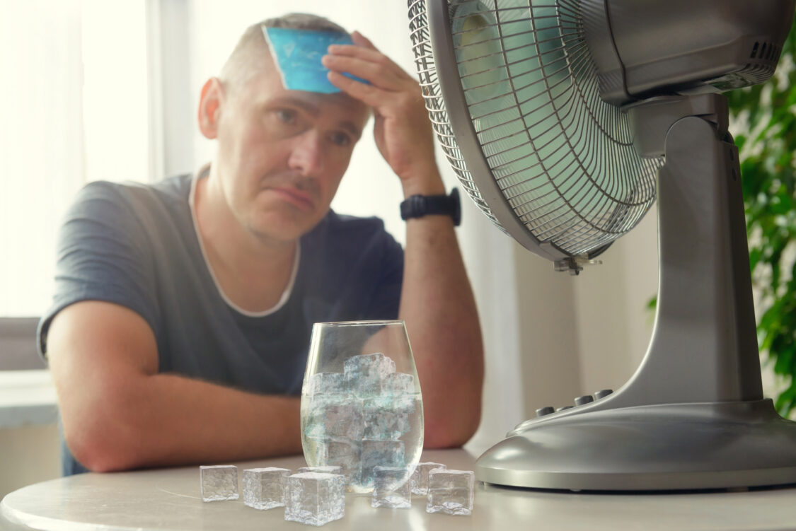 man suffering from the heat sitting in front of a fan and a glass of water 