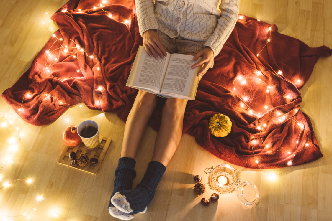 Girl sitting on a blanket and reading book with a cup of coffee to go wearing woolen sweater and knitted shorts - home autumn cosiness and comfort concept, top view