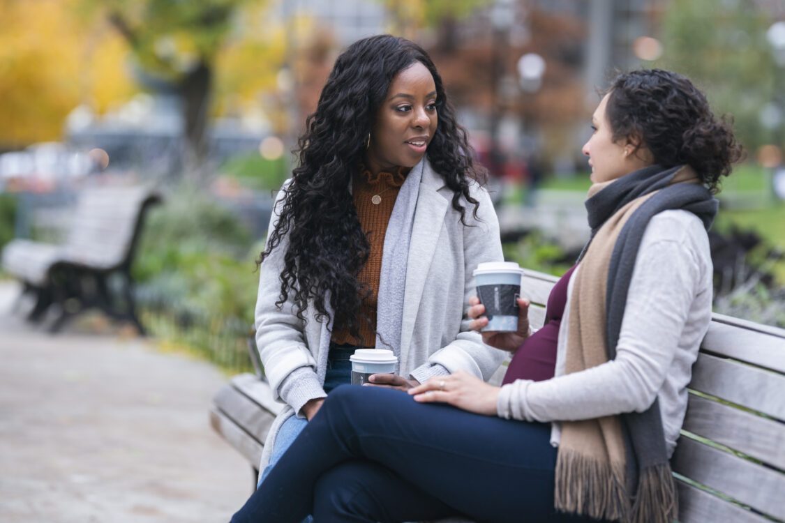 Pregnant woman talking with female friend while relaxing on park bench