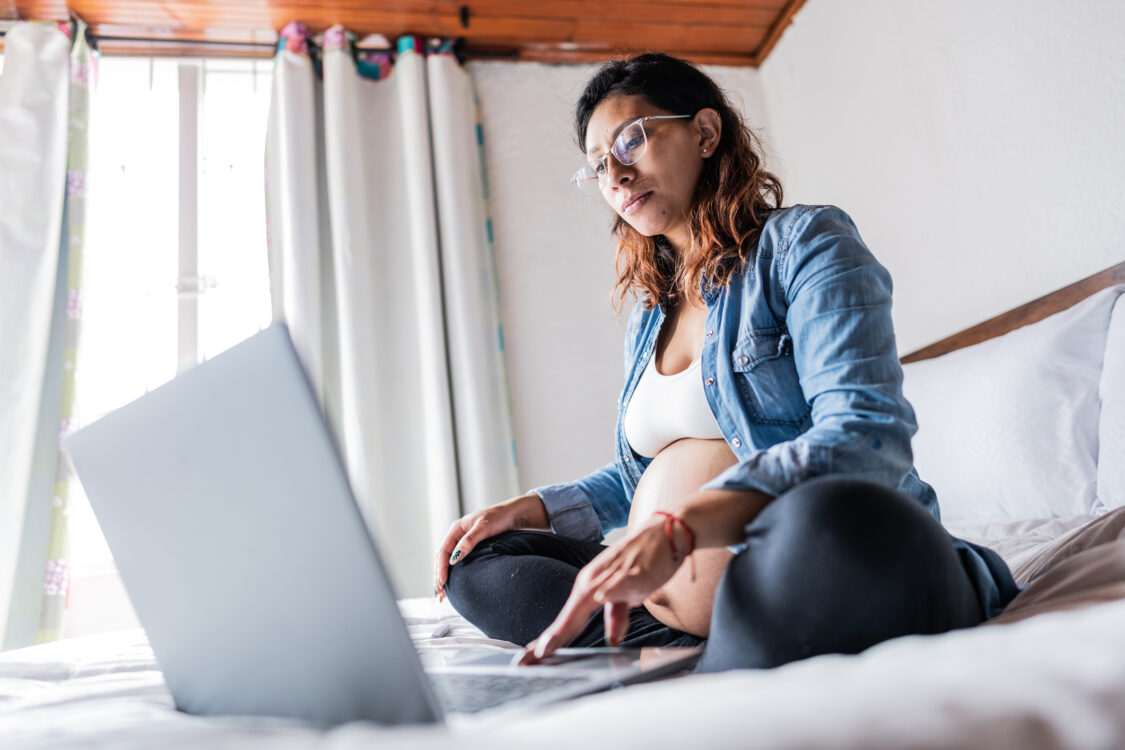 Pregnant woman using laptop sitting on bed at home