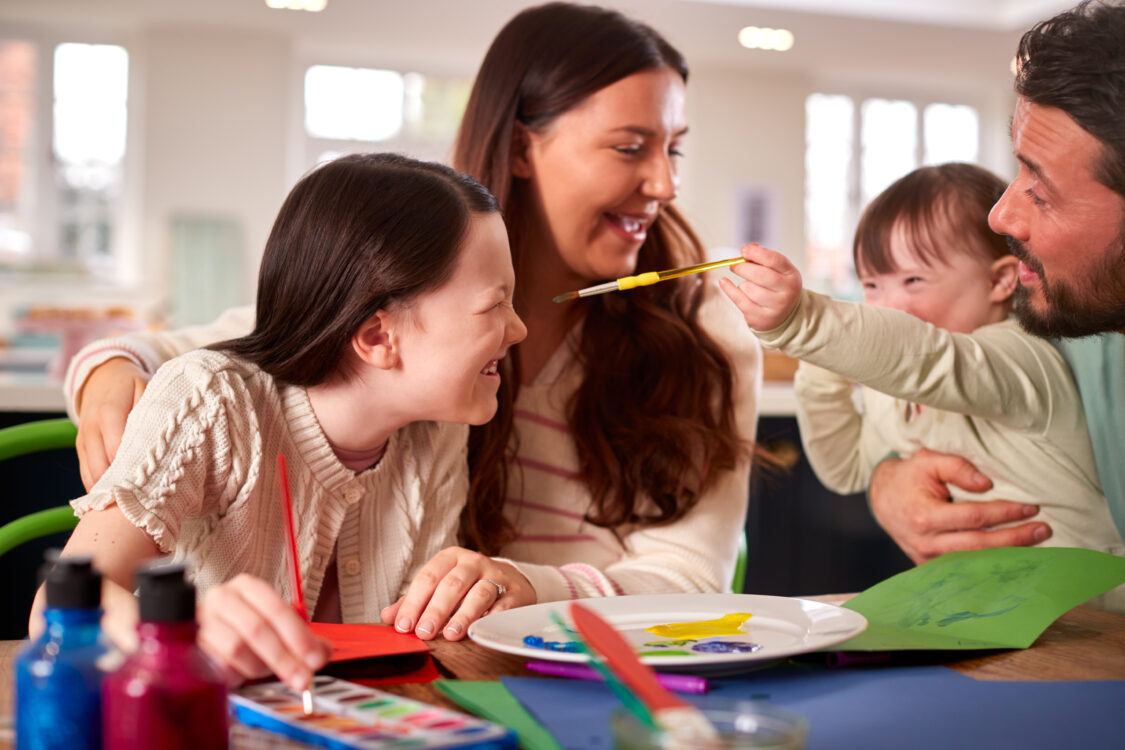 Family With Down Syndrome Daughter Sitting Around Table At Home Doing Craft Together