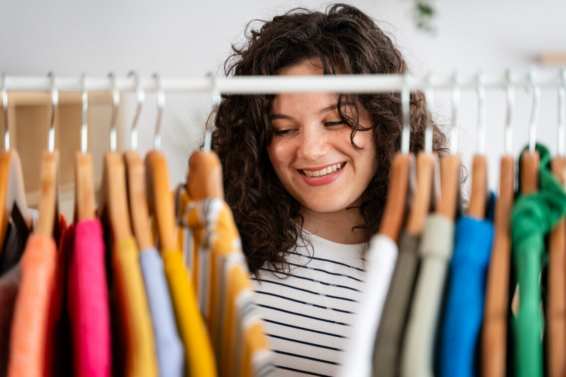 Young woman with curly hair smiling while sifting through a variety of brightly colored clothing on a white wardrobe rack